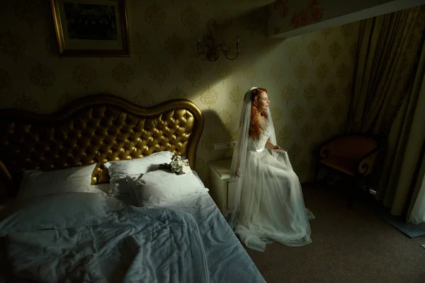 Hermosa señora pelirroja sexy en elegante vestido de novia blanco. Retrato de moda de modelo en interiores. Mujer bella sentada cerca de su cama — Foto de Stock