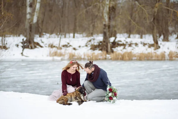 Elegante hermosa pareja joven novia y novio, perro sentado en el fondo de un río paisaje de invierno . — Foto de Stock