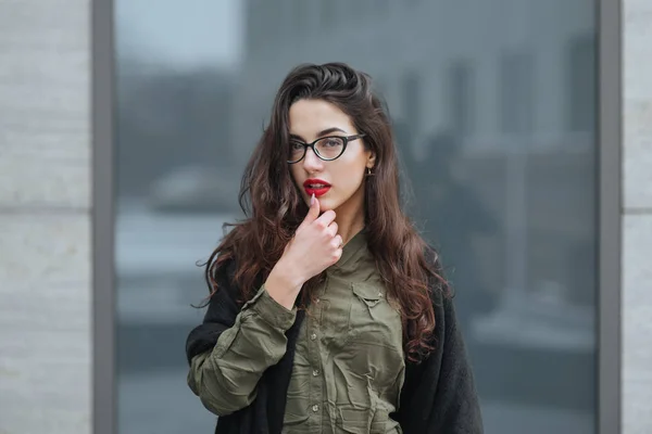 Fashion consept: beatiful young girl with long hair, glasses, red lips standing near modern wall wearing in green suit and grey jeans. She wanted talking on phone. — Stock Photo, Image