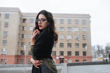 Moda consept: güzel uzun saçlı genç kız gözlük, yeşil takım ve gri kot modern duvarının yakınında duran kırmızı dudaklar. Evet istedi.