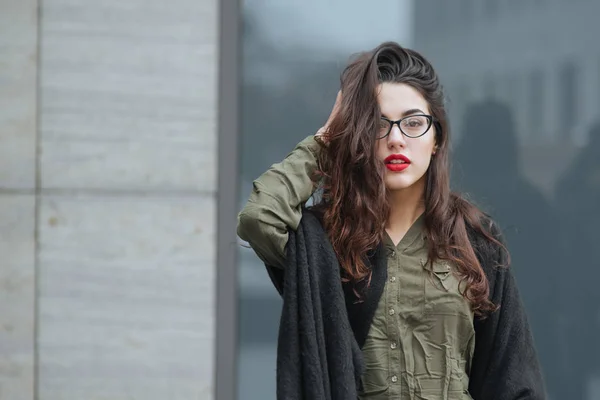 Fashion consept: beatiful young girl with long hair, glasses, red lips standing near modern wall wearing in green suit and grey jeans. She wanted talking on phone.