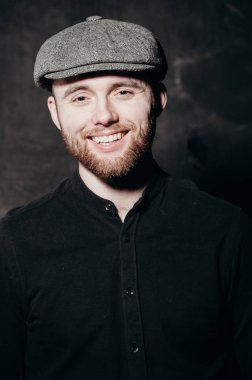 Handsome young bearded man in hat on grey background looking at camera. Portrait of laughing young man against grey wall. Happy guy smiling.