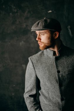 Serious handsome young bearded man in hat on grey background looking at side. Portrait of man against grey wall.