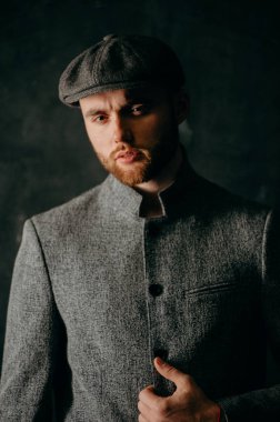 Serious handsome young bearded man in hat on grey background looking at camera. Portrait of man against grey wall.