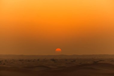 Dubai çölünün üzerinde canlı, turuncu bir günbatımı. Alevli güneş ufka doğru alçalır, sıcak, atmosferik bir parıltı yayar, kayan kumullar ve puslu manzaralar boyunca..