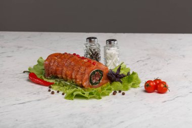 Çiğ, doldurulmuş, kırmızı baharatlı domuz eti rulosu, taze marul yapraklarının üzerine yerleştirilmiş. Ispanak dolgulu pişirilmemiş et Roulade, kiraz domatesli beyaz mermer bir masaya yerleştirilmiş kırmızı biber, mor fesleğen ve tuz ve biber..