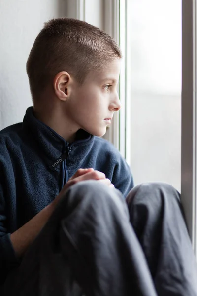 Eenzame verweesde jongen zit aan de glazen raam — Stockfoto