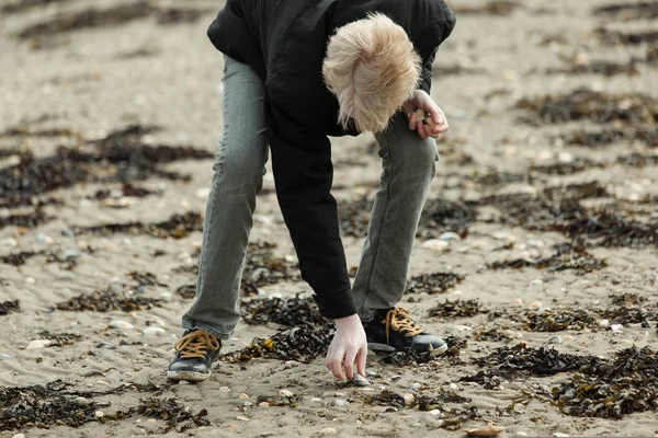 Blond boy plocka upp sten på stranden — Stockfoto