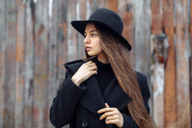 Siyah şapkalı, güzel, modaya uygun, uzun saçlı, odunsu arka planda poz veren bir kadın.