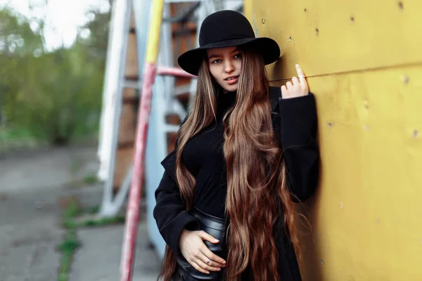 Junge schöne modische Frau mit schwarzem Hut und langen Haaren posiert auf verschwommenem Hintergrund — Stockfoto