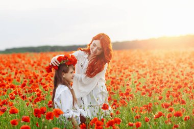 Beyaz elbiseli kızıl saçlı küçük mutlu kız yaz günbatımında gelincik tarlasında çelenk yapıyor.