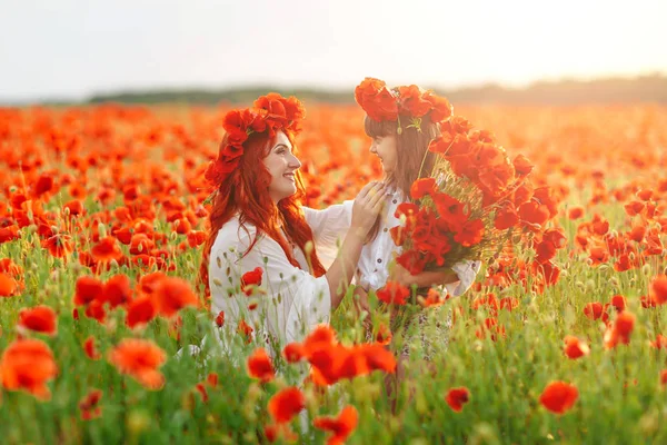 Beyaz elbiseli ve çelenkli kızıl saçlı küçük bir kız sıcak yaz günbatımında haşhaş tarlasında haşhaş buketi ile poz veriyor.