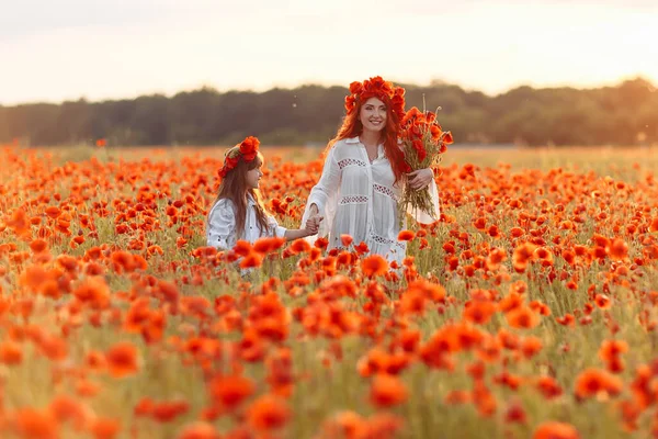 Little girl with redhead mother in white dresses and wreathes walking with bouquet of poppies on poppy field at warm summer sunset