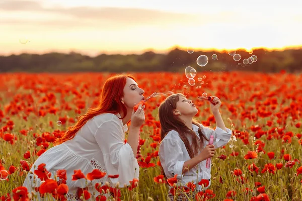 Little happy girl with redhead mother in white dresses blow bubbles on poppy field at warm summer sunset