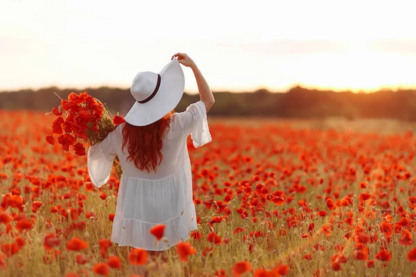 Beautiful redhead woman in hat on green field with bouquet of poppies in summer sunset