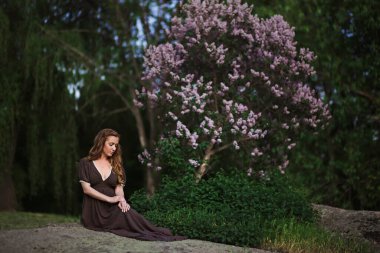 Güzel rüya gören kızıl saçlı genç kadın baharda çiçek açan leylağın uzun elbisesiyle poz veriyor.