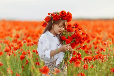 Beyaz elbiseli küçük tatlı kız elinde bir buket gelincik ile tarla gelinciği çelengi oynuyor.