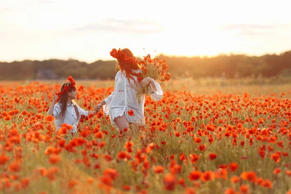 Little girl with redhead mother in white dresses and wreathes walking with bouquet of poppies on poppy field at summer sunset