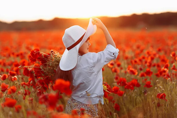 Pequena menina bonito em vestido branco e chapéu branco posando no campo de papoilas com um buquê de papoilas em suas mãos no pôr do sol de verão — Fotografia de Stock