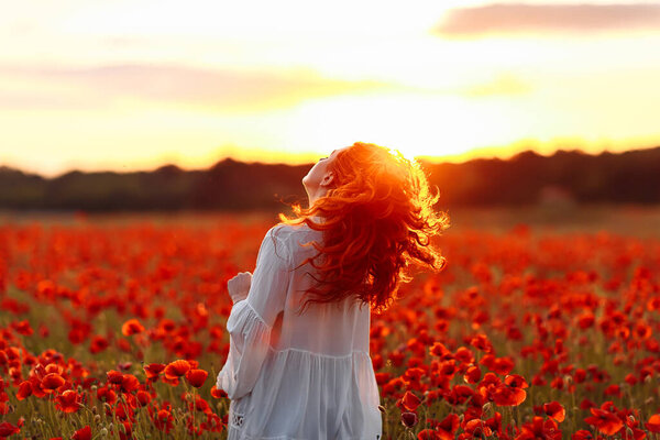 Happy redhead smiling woman in white dress on field of poppies at warm summer sunset