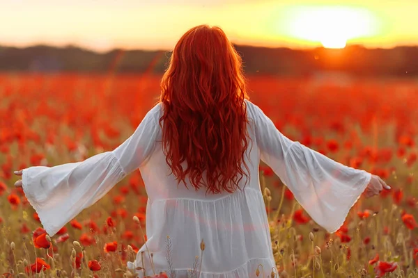 Happy redhead smiling woman in white dress on field of poppies at summer sunset