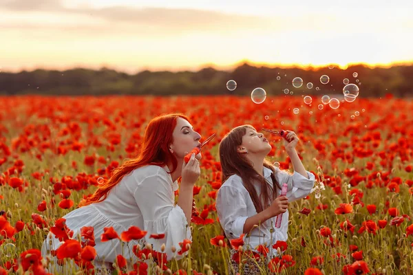 Petite fille avec mère rousse en robes blanches soufflent des bulles sur le champ de pavot au coucher du soleil d'été — Photo