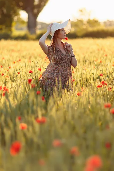 Beautiful redhead woman in white hat posing on green field with poppies