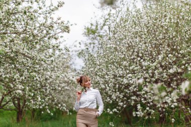 Beyaz tişörtlü güzel sarışın kadın çiçek açmış elma ağacının altında poz veriyor ve bahar bahçesinde yeşil çimenler.