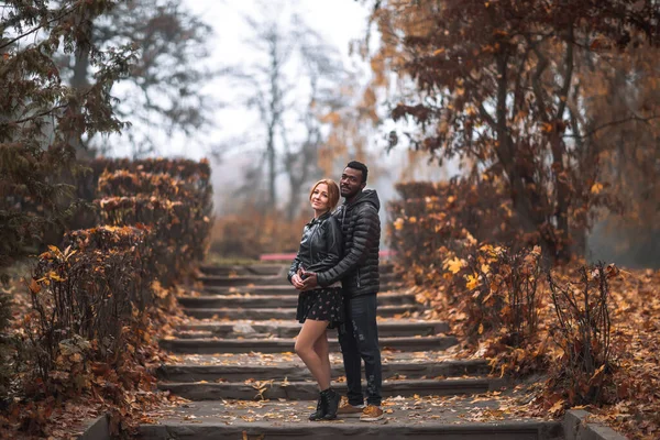Interracial couple posing on stairs in blurry autumn park background, black man and white redhead woman