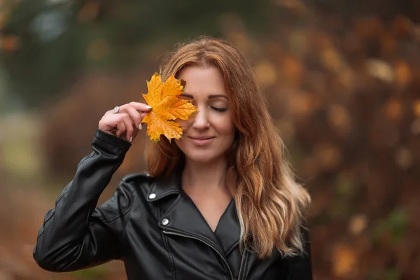 Portrait of smiling redhead woman with yellow leaf with long curly hair on blurred autumn background. Girl on fabulous background of park with orange autumn leaves. and road.