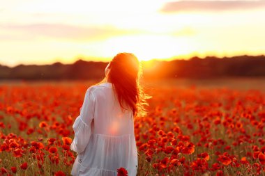 Sıcak yaz günbatımında haşhaş tarlasında beyaz elbiseli mutlu kızıl saçlı kadın.