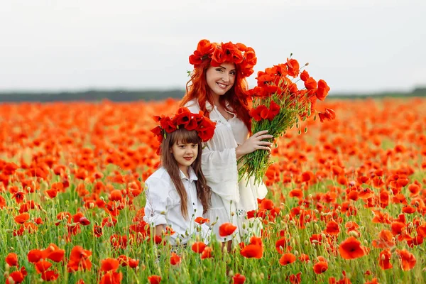 Little girl with redhead mother in white dresses and wreathes poses with bouquet of poppies on poppy field at warm summer sunset