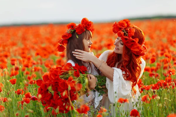 Little happy girl with redhead mother in white dresses makes wreath on poppy field at warm summer sunset