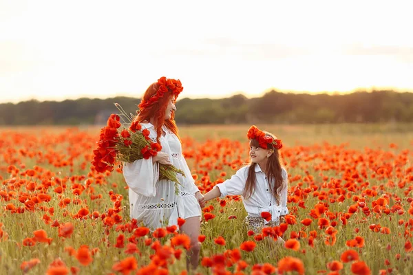 Little girl with redhead mother in white dresses and wreathes walking with bouquet of poppies on poppy field at warm summer sunset