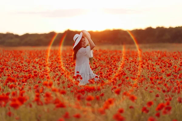 Beautiful redhead woman in hat on green field with bouquet of poppies in summer sunset