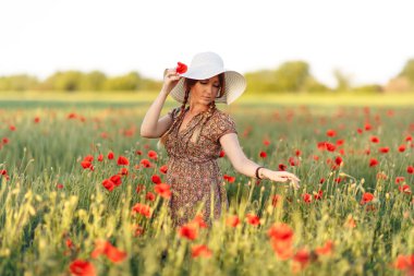 Beyaz şapkalı güzel kızıl kadın yeşil tarlada gelinciklerle poz veriyor.