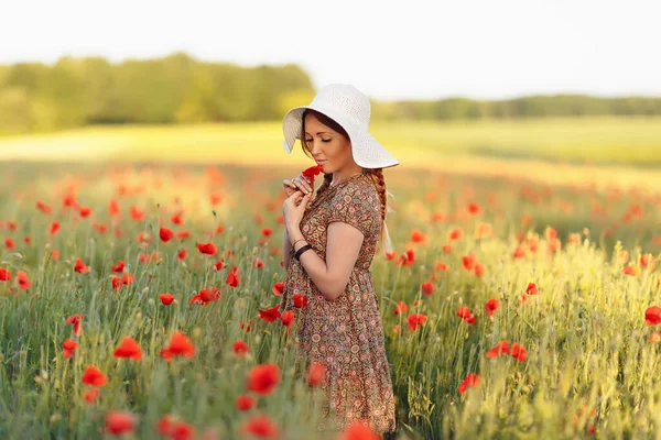 Beautiful redhead woman in white hat posing on green field with poppies