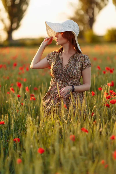 Redhead woman in white hat on green field with poppies in summer sunset