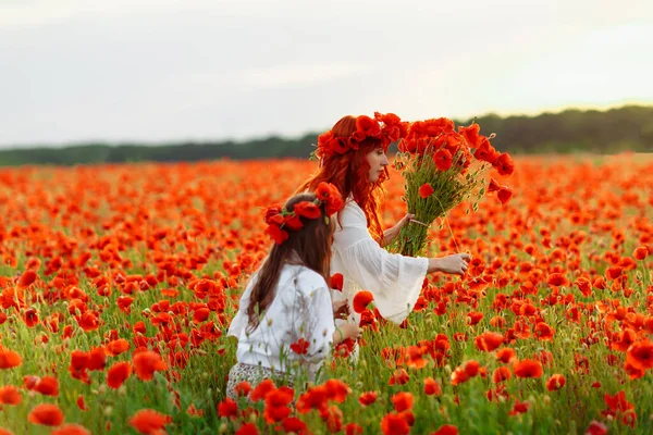 Little girl with redhead mother in white dresses and wreathes poses with bouquet of poppies at warm summer sunset on poppy field