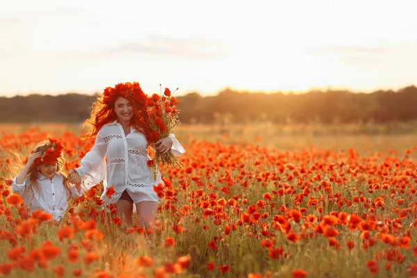 Little girl with redhead mother in white dresses and wreathes walking at warm summer sunset with bouquet of poppies on poppy field
