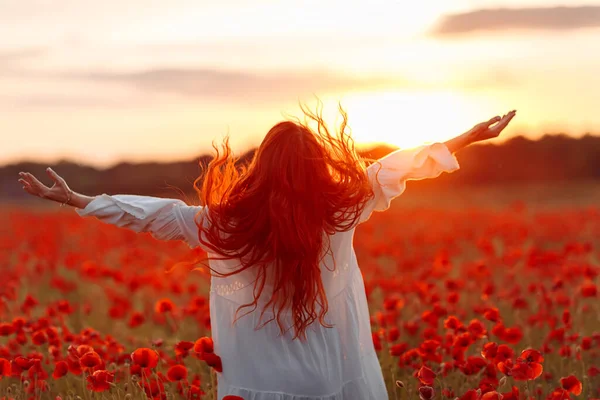 Redhead happy woman in white dress on field of poppies at warm summer sunset