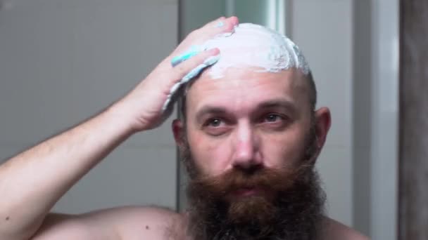 Τολμηρός μεσήλικας γενειοφόρος άντρας βάζει αφρό ή τζελ ξυρίσματος στο κεφάλι του κοιτάζοντας στον καθρέφτη στο μπάνιο.. — Αρχείο Βίντεο