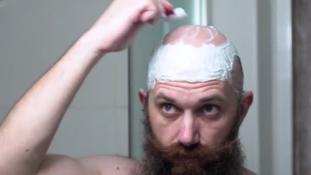 El hombre barbudo se afeita la cabeza mirando en el espejo en el baño después de la ducha. Acelerar vídeo . — Vídeo de stock