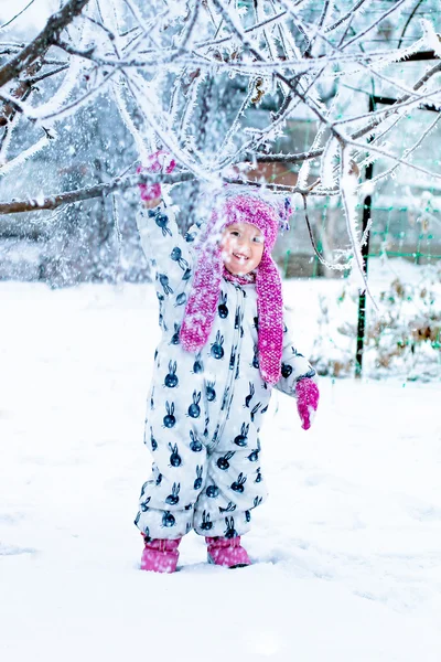 Enfant par temps neigeux. La neige tombe de l'arbre. Bébé fille en suite blanche et chapeau rose, bottes gants dans le parc d'hiver. Heureux . — Photo