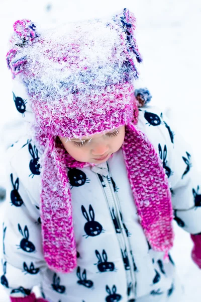 Enfant par temps neigeux. Portrait de bébé fille en costume de neige blanche et chapeau rose dans le parc d'hiver. dans  . — Photo