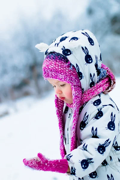 Enfant par temps neigeux. Bébé fille en costume de neige blanc et chapeau rose regardant sur les gants avec de la neige . — Photo