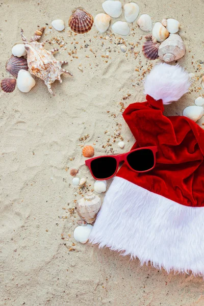 Boże Narodzenie kapelusz i czerwone okulary na plaży. Santa okulary piasek w pobliżu pociski. Holiday. Nowy rok wakacje. Kopiować miejsca. Ramki. Widok z góry. — Zdjęcie stockowe
