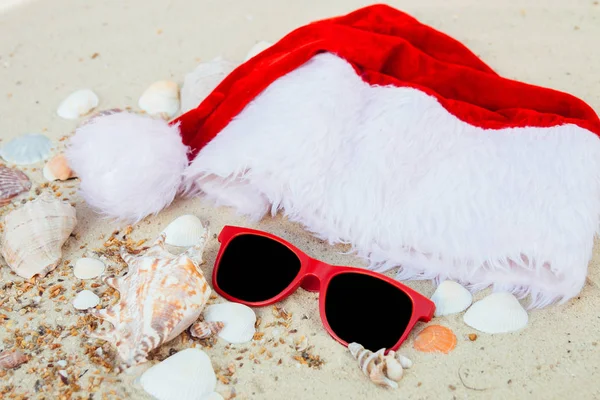 Boże Narodzenie kapelusz i czerwone okulary na plaży. Santa okulary piasek w pobliżu pociski. Holiday. Nowy rok wakacje. Kopiować miejsca. Ramki. Widok z góry. — Zdjęcie stockowe