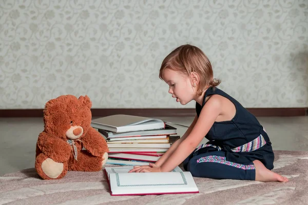 Bambina in abito blu scuro libro lettura seduta sul pavimento vicino orsacchiotto. Bambino legge storia per giocattolo . — Foto Stock