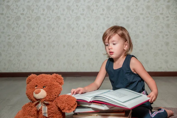 테 디 베어 근처 바닥에 앉아 어두운 파란색 드레스 읽기 책에 작은 소녀. 어린이는 장난감에 대 한 이야기를 읽으십시오. — 스톡 사진
